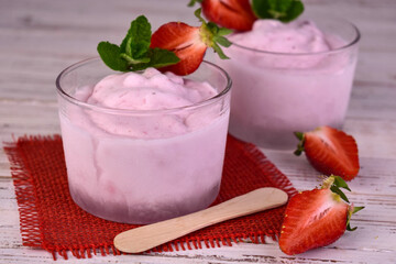 Two servings of strawberry ice cream in glasses on a white wooden background.
