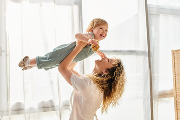 A curly mother joyfully lifts her toddler daughter up into the air, expressing love and playfulness...