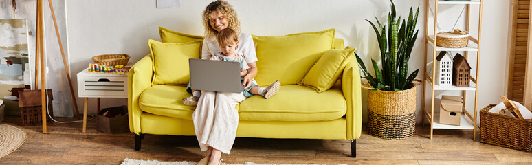 A woman with curly hair is seated on a yellow couch, focused on her laptop.