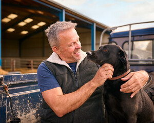 Mature Male Farm Worker Sitting On Tailgate Of Off Road Farm Vehicle With Dog