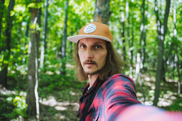 A man with long hair in a plaid shirt takes a selfie in a forest. The dense trees and sunlight...