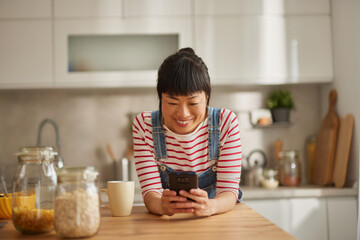 Beautiful Asian woman using mobile phone while drinking coffee in the kitchen