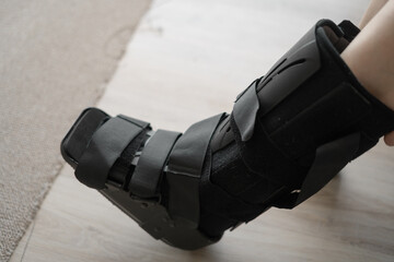 Close-up of female leg with black splint, walker boot, orthosis, sprained ankle at home. Ankle...