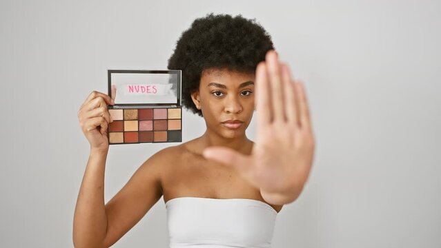 Confident african american woman with serious expression, halting makeup misuse with firm stop gesture, displaying protective defense over stark isolated white background