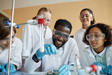 Cheerful African American teacher and group of ethnically diverse kids doing lab experiment during...