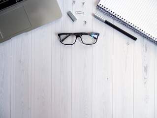 Laptop, notatnik, okulary, długopis, spinasz, pinezki i zszywki ułożone symetrycznie na białym biurku. © KJP Photography