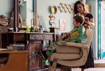 A happy young boy is having his hair cut by a stylist in a charming, vintage-themed salon filled...