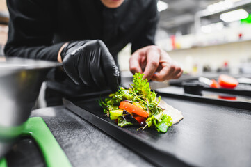 A chef’s hands, gloved in black, meticulously garnishing a dish with fresh herbs and vibrant...