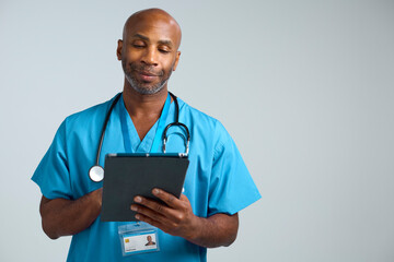 Studio Shot Of Mature Doctor Wearing Scrubs Using Digital Tablet Against White Background