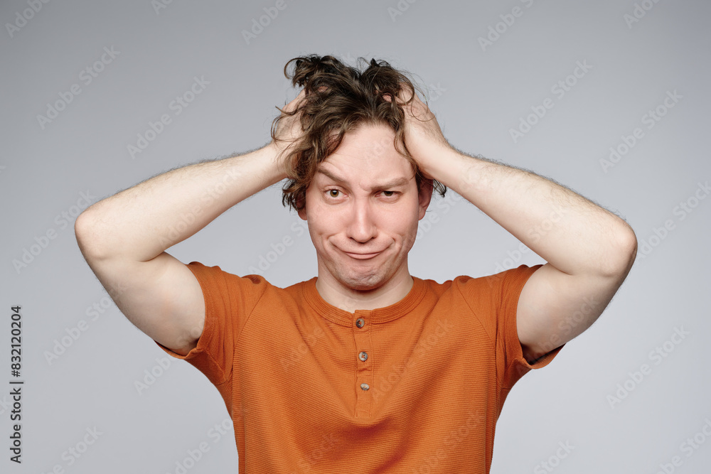 Wall mural Studio portrait of confused young Caucasian man wearing orange t-shirt posing for camera with both his hands on head