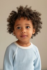 Naklejka premium young African-American child with short curly hair, wearing a light blue waffle-knit top and matching pants, standing in a minimalist studio setting with a soft beige background