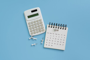 Top view of white calendar, calculator and thumbtack on blue background
