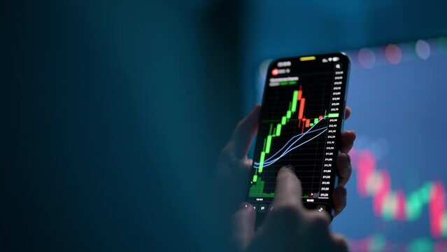 Tracking shot of female stock trader checking stock market data using mobile phone, scrolling, touching stock market chart on touch screen device, blurred computer monitor with chart of background.