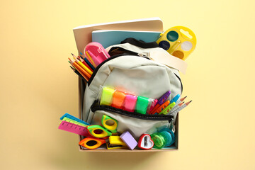 Open backpack filled with colorful school supplies such as pencils, markers, scissors, and notebooks in donation box on a pastel yellow background. Donation concept.