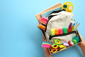 Womens hands holding donation box with backpack full of vibrant school supplies on blue background....