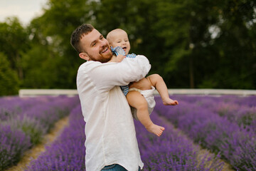 A bearded man in a white shirt is lifting baby in air on a lavender field. Daddy is lifting up daughter and have fun. 