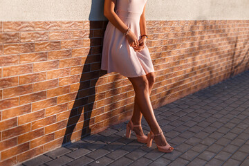 beautiful young girl in a fashion pink dress with heels stands near the wall on a summer day at sunset
