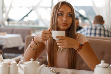 Beautiful fresh tender young woman in a fashion pink dress sits in a summer cafe and drinks coffee