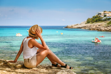 A woman in a straw hat, sitting by the beach, looking out at the clear blue sea, highlighting a...