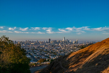 The image shows a panoramic view of a city skyline with numerous buildings under a clear blue sky, taken from a high vantage point with a hill in the foreground.