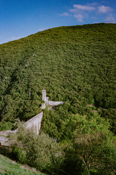 A verdant hillside with dense foliage hosts an old stone structure, likely a historical building, nestled among the greenery under a clear blue sky.