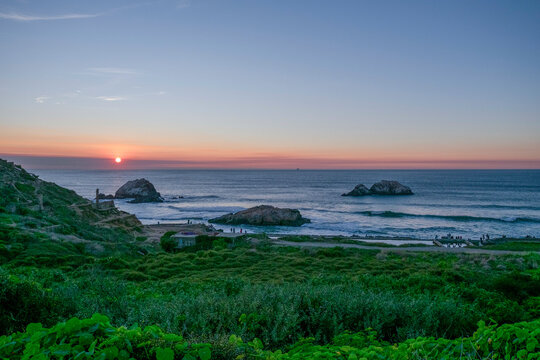 Sunset view over a coastal landscape with undulating hills, lush greenery in the foreground, and several rock formations in the ocean. - Powered by Adobe
