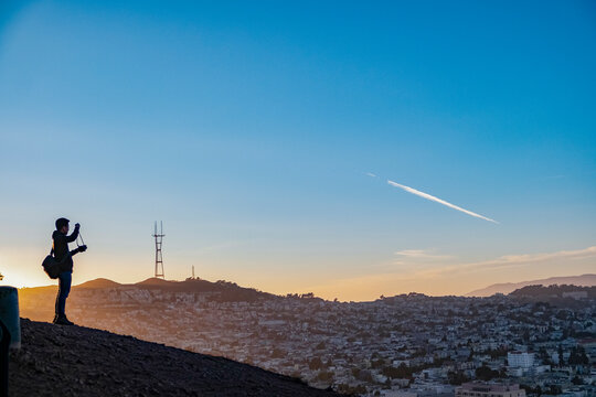 A person stands on a hill during sunset, taking a photograph of the cityscape with a tower in the distance. - Powered by Adobe