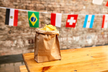 A paper bag filled with potato chips is placed on a wooden table with international flags hanging in the background.
