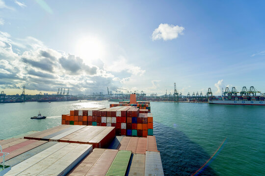 A cargo ship is laden with colorful containers under a partly cloudy sky, sailing near a busy port with numerous cranes and shipping infrastructure.