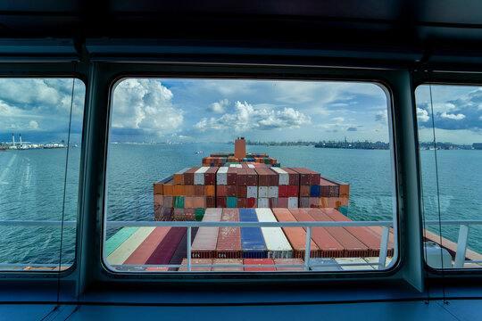 A view from a ship's bridge window overlooking a large stack of colorful shipping containers on the vessel's deck, with the ocean and cloudy sky in the background.