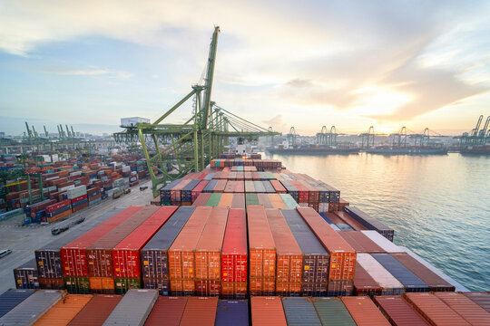 A birds-eye view of a cargo ship loaded with colorful containers docked at a busy port during sunset. - Powered by Adobe