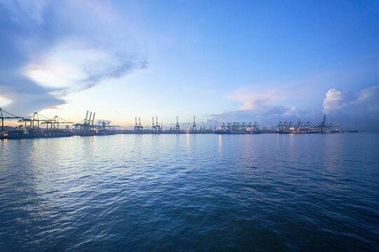 Dawn breaks over a busy port with rows of cranes lining the docks, and container handling operations underway, against a backdrop of a calm sea and a clear sky with soft clouds.
