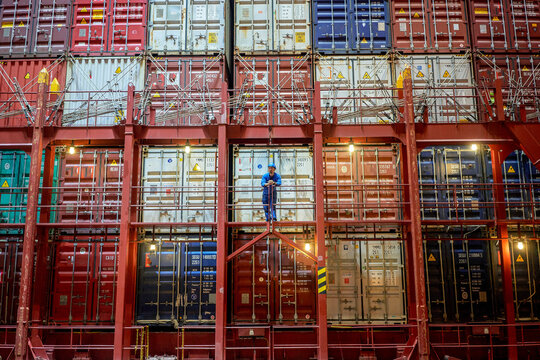 A worker stands on a metal stairway amidst a large stack of colorful shipping containers at night, illuminated by ambient lights.