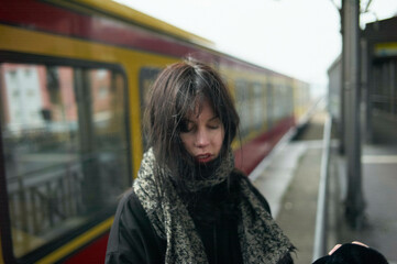 A woman stands on a train platform, her face partially obscured by windswept hair, as she appears deep in thought.