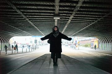 An individual is standing in the center of a tunnel-like structure with their arms outstretched, eyes closed, and a serene expression. The open end of the tunnel leads to a bright cityscape.