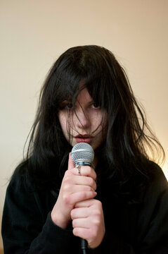 A person with messy black hair covers part of their face, holding a microphone close to their lips, in front of a plain beige background.