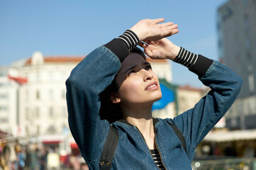 A young woman is outdoors on a sunny day, shading her eyes with her hand as she looks upward, with a city street and buildings in the background.