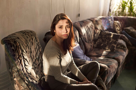A young woman is sitting on a floral couch, bathed in natural sunlight, with a pensive expression on her face.