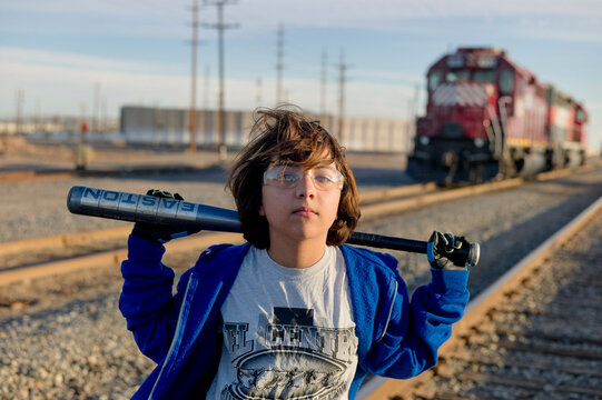 A kid stands on a railway track holding a baseball bat on their shoulder, with a train in the background during the daytime.
