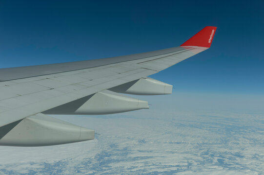 An airplane wing with a red tip soars above the clouds and ice-covered landscape below, capturing the majesty of flight in a clear blue sky.