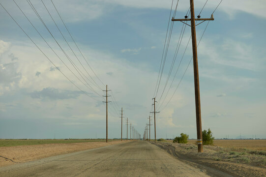 A dirt road stretches into the distance flanked by parallel rows of electrical poles under a vast cloudy sky.