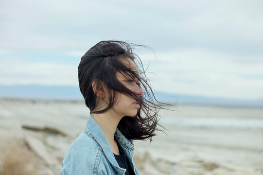 A woman stands on a beach with her hair blowing in the wind, looking away from the camera.