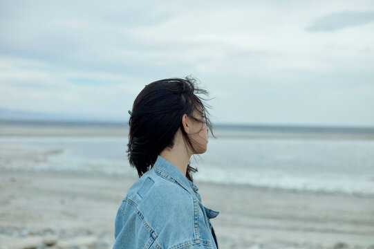A profile view of a person with black hair wearing glasses and a denim jacket, standing in front of a serene beachscape and overcast sky.
