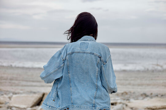 A person with long dark hair wearing a denim jacket stands facing a tranquil sea, with the horizon in the background under an overcast sky.