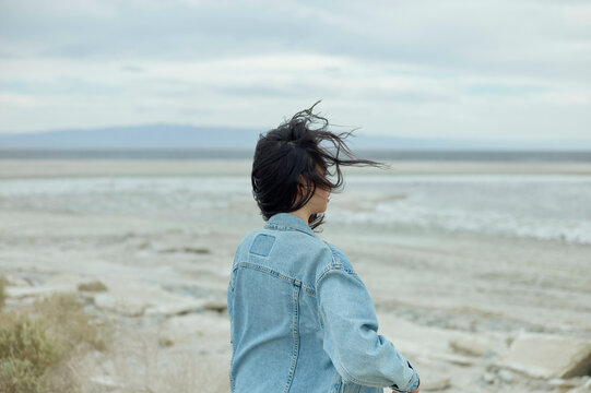 A person in a denim jacket stands on a shore with their hair blowing in the wind, gazing out over a calm body of water under an overcast sky.