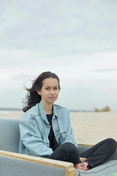 A woman sits on a beach chair with her legs crossed, wearing a denim jacket and looking directly at the camera with a subtle smile, against a cloudy sky.