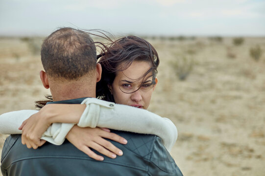 A woman embraces a man from behind in a barren landscape, her expression a mix of pensiveness and concern.
