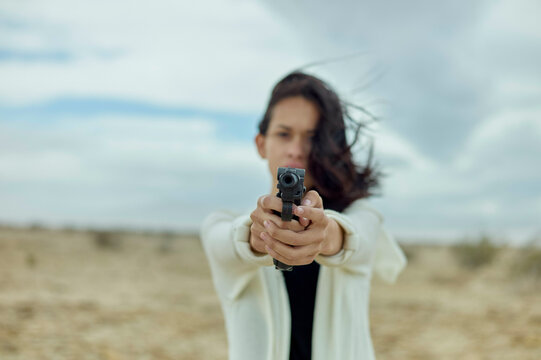 A woman with her hair blowing in the wind is holding a gun aimed forward with a focused expression, standing in a barren landscape under a cloudy sky.