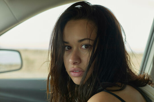 A woman sits in a car, turning to look at the camera with an intense gaze, her dark hair tousled and a deliberate, pensive expression on her face.