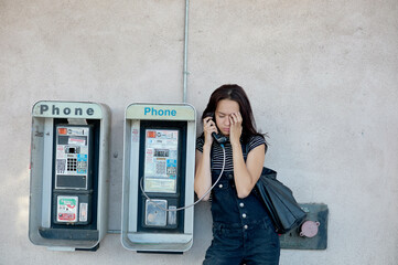 A woman is standing between two public payphones, engaged in a conversation on one of them, with her eyes closed and a slight frown, possibly indicating a serious or troubling discussion.